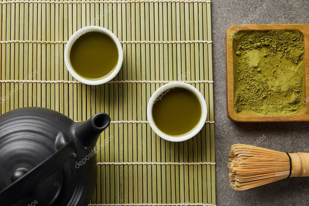 top view of white cups with green tea near black teapot on bamboo table mat with matcha powder on wooden board